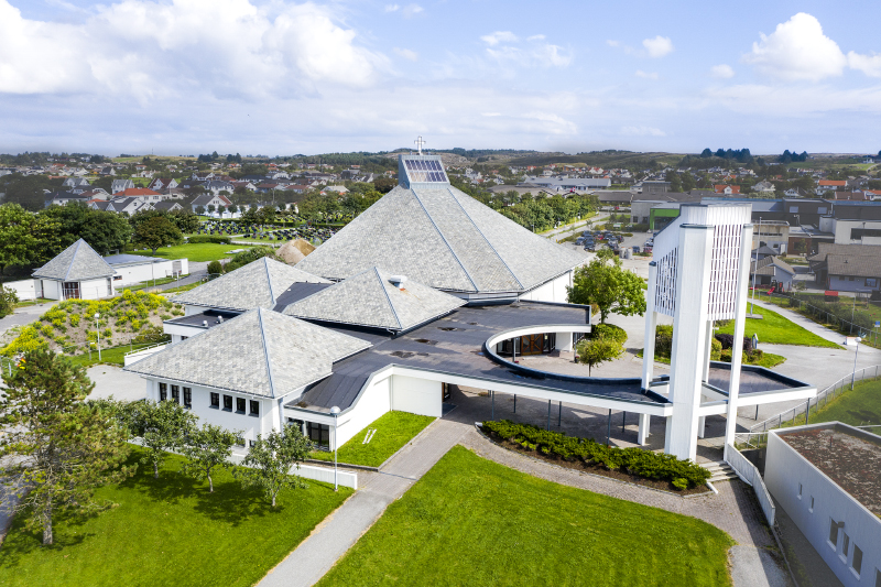 Stor moderne kirke med mange takflater og frittstående klokketårn, sentralt plassert i byområdet Åkrehamn. Dronebilde.
