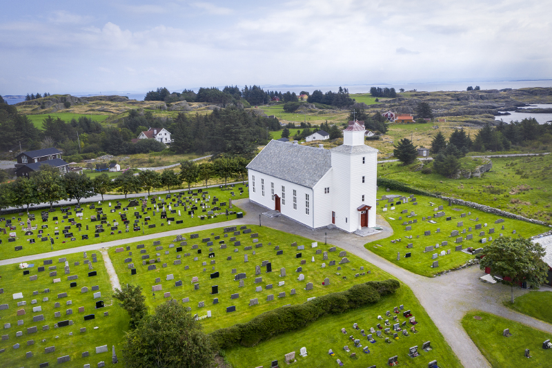 Hvit trekirke med gravplass rundt, omgitt av trær og kystlandskap nær Skudeneshavn. Dronebilde.
