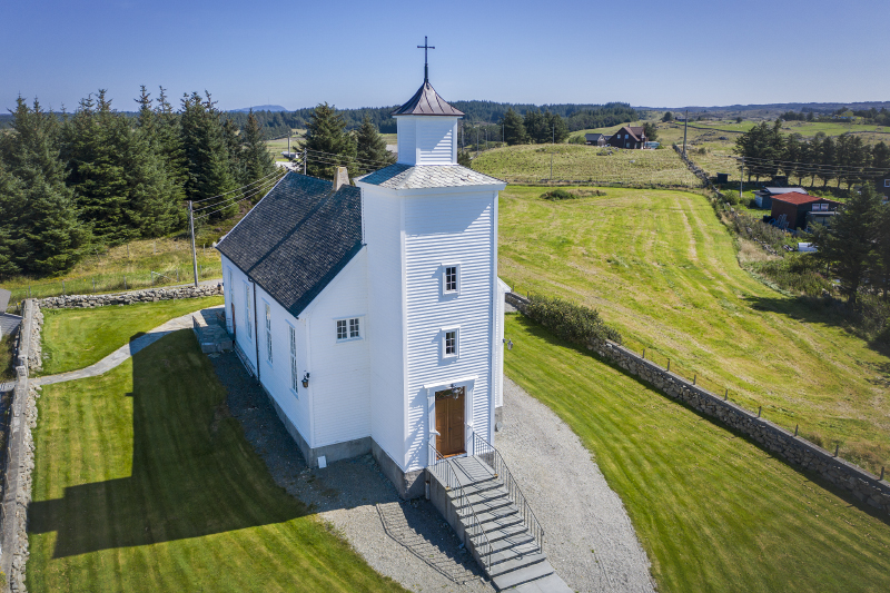 Liten hvitmalt trekirke på en åpen gresslette med skog i bakgrunnen. Dronebilde.
