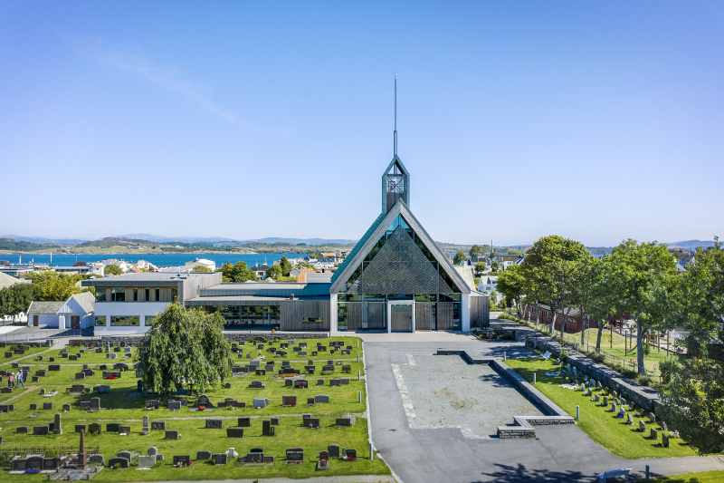 Moderne kirke med spisst tårn og trekantet glassfasade, omgitt av gravplass og bymiljø. Dronebilde.
