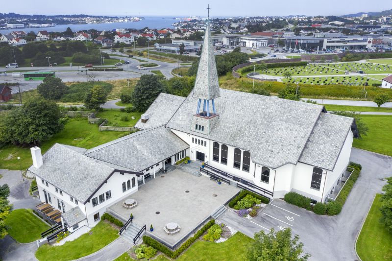 Stor hvit kirke med høyt spir og tilknyttede bygninger, beliggende i et urbant område med utsikt mot sjøen. Dronebilde.