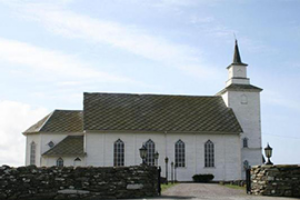 Tradisjonell hvitmalt trekirke med spisst tårn og gamle steingjerder rundt, i landlige omgivelser. Foto.