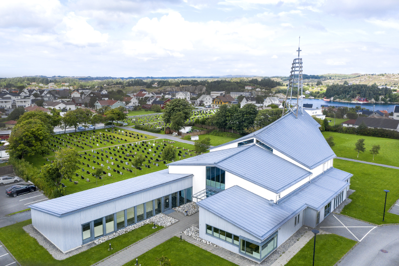 Moderne kirke med blått tak og glasspartier, omgitt av en gravplass og boligområder. Dronebilde.
