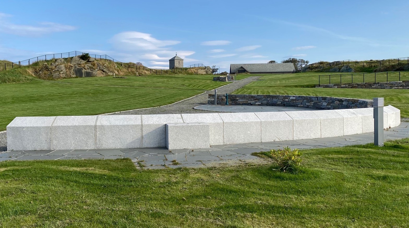 En navnet minnelund med en rund steinmur og lyse granittblokker i forgrunnen. Området er omgitt av velholdt grønt gress, med en steinlagt sti som leder opp til en liten bygning med spisst tak i bakgrunnen. Blå himmel med lette skyer gir en fredelig atmosfære. Foto.