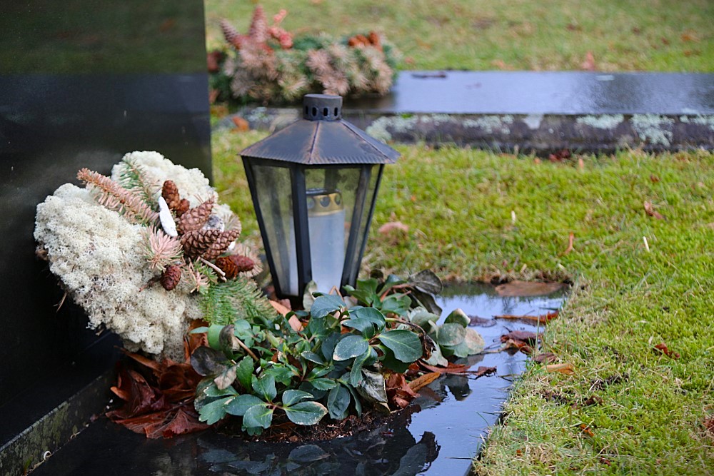 En grav med en svart gravstein, pyntet med en lykt, vintergrønn beplantning og en dekorasjon av lav, kongler og granbar. Gresset rundt er vått, og det ligger noen visne blader på bakken, noe som gir en høstlig eller vinterlig stemning. Foto.